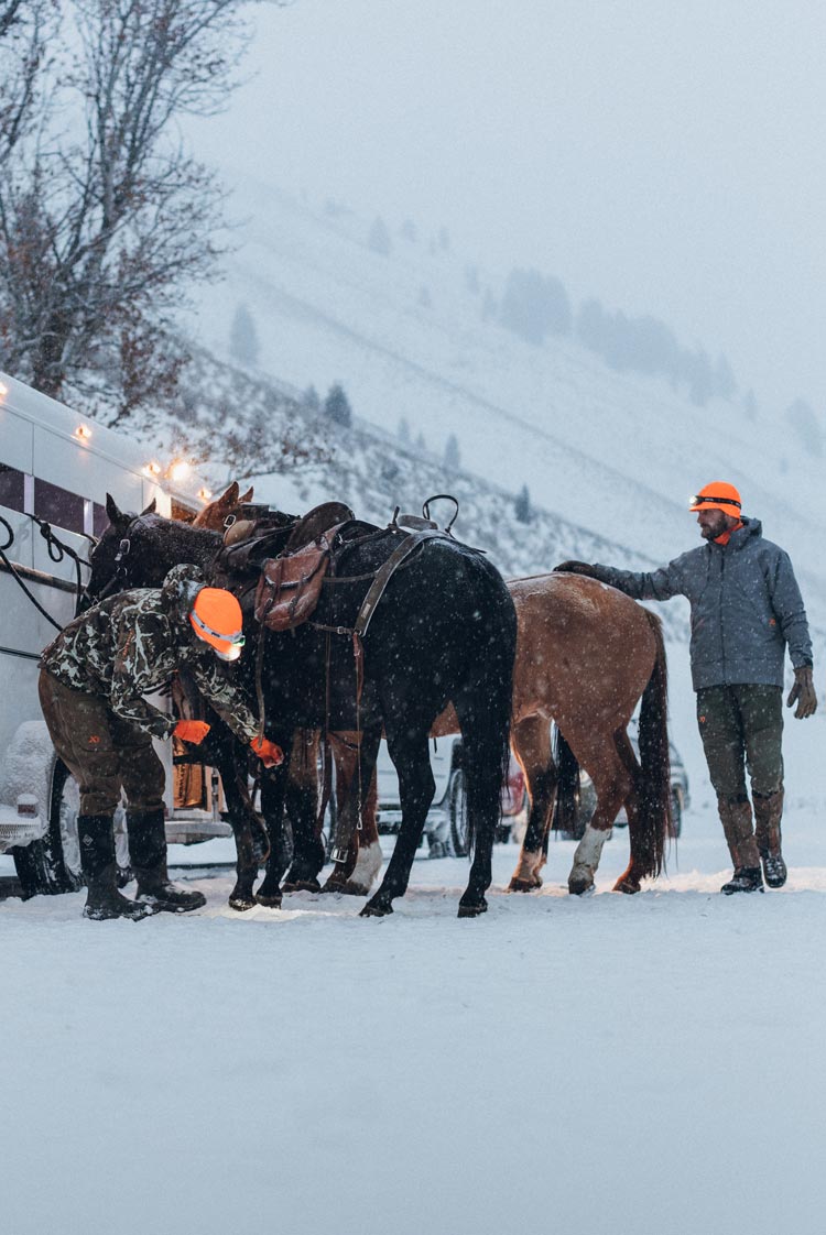 Two hunters in heavy jackets and cold weather gear place saddles on a group of horses tethered to a trailer.