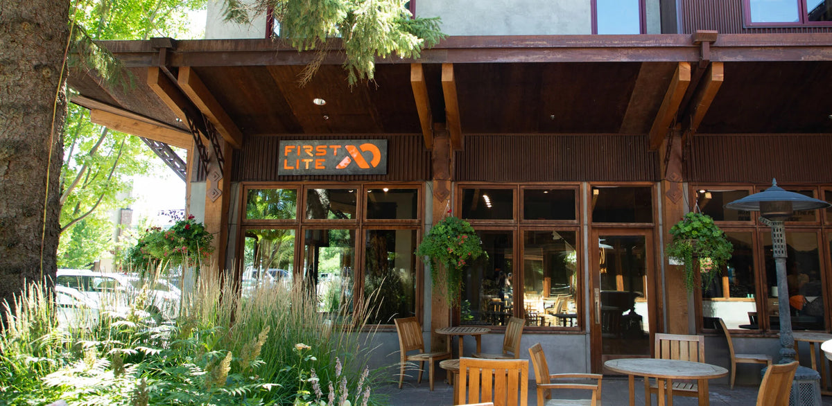 Rustic brown "FIRST LITE" store with orange sign, large windows, outdoor patio, and plants.