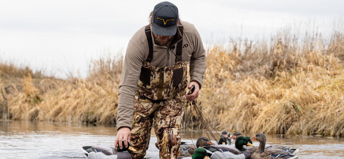 Man in tan jacket and camo waders setting duck decoys in water.