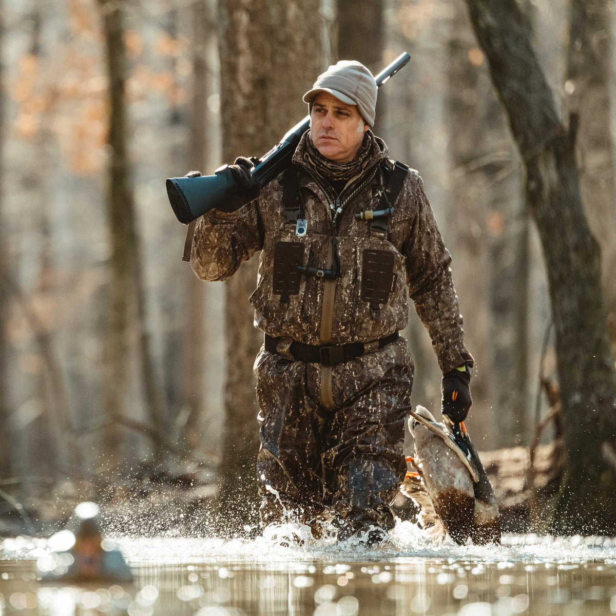 Man in brown camouflage hunting waders walks through shallow water carrying a shotgun and two ducks.