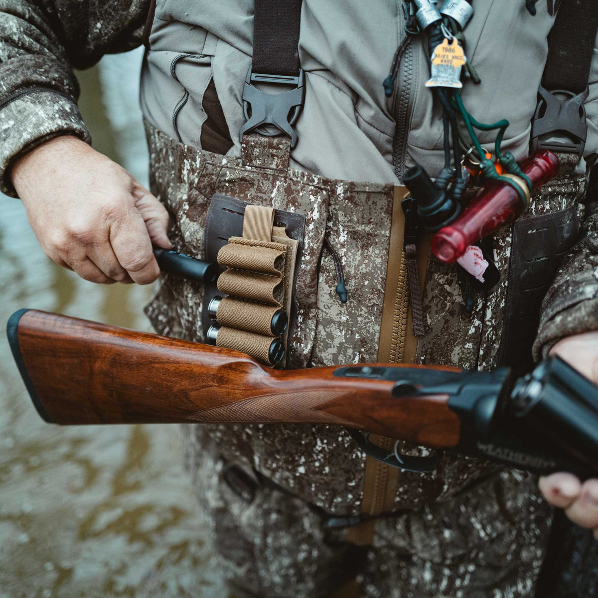 A person in camouflage waders loads a shotgun shell into a dark wood shotgun, with a tan shell holder secured to their chest.