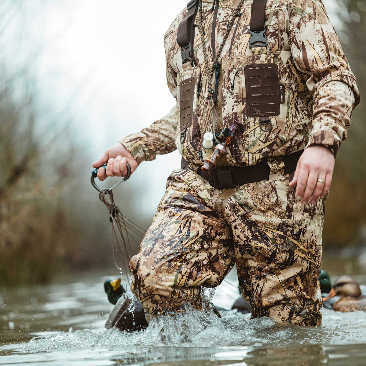 Brown camouflage hunting waders and jacket worn by a person standing in splashing water, holding a decoy line with duck calls on a chest lanyard.