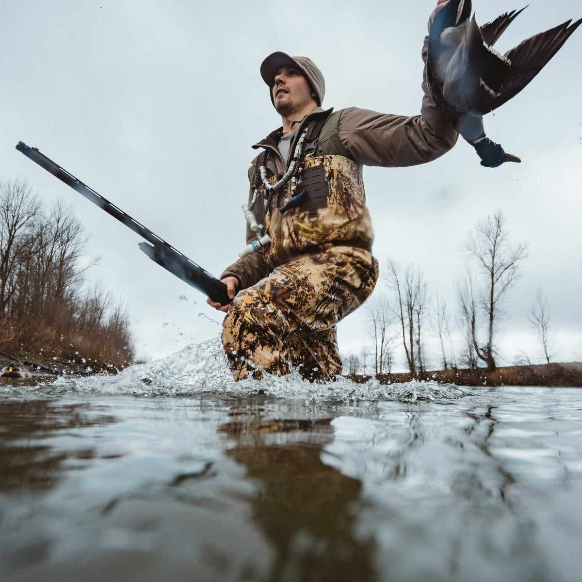 A duck hunter in brown and green camouflage waders and a brown jacket splashes through water, holding a black shotgun and a dark duck.