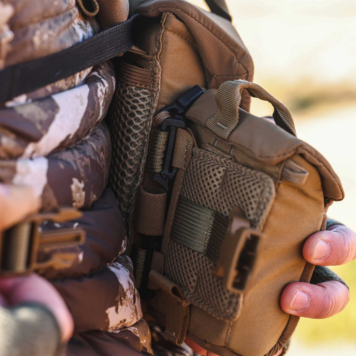 Coyote brown tactical gear pouch with mesh pockets, webbing, and a black buckle, held by a person in camouflage.