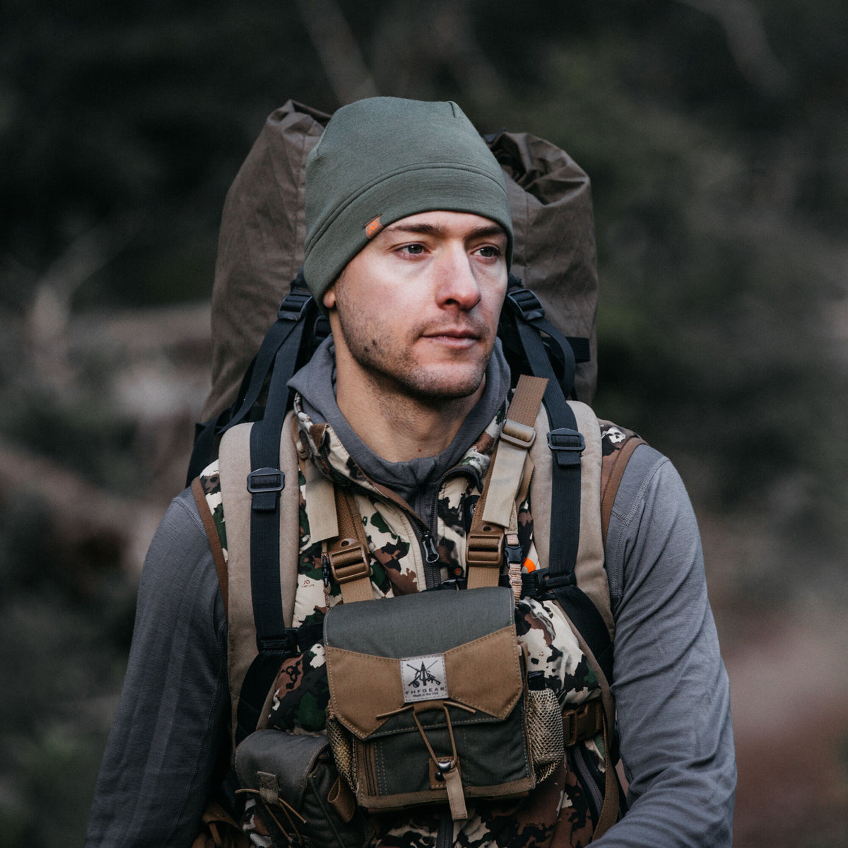 An olive green beanie with a small orange tag, worn by a man outdoors.