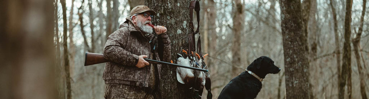 Camo-clad hunter blows red duck call, with shotgun, retrieved ducks, and black Lab.