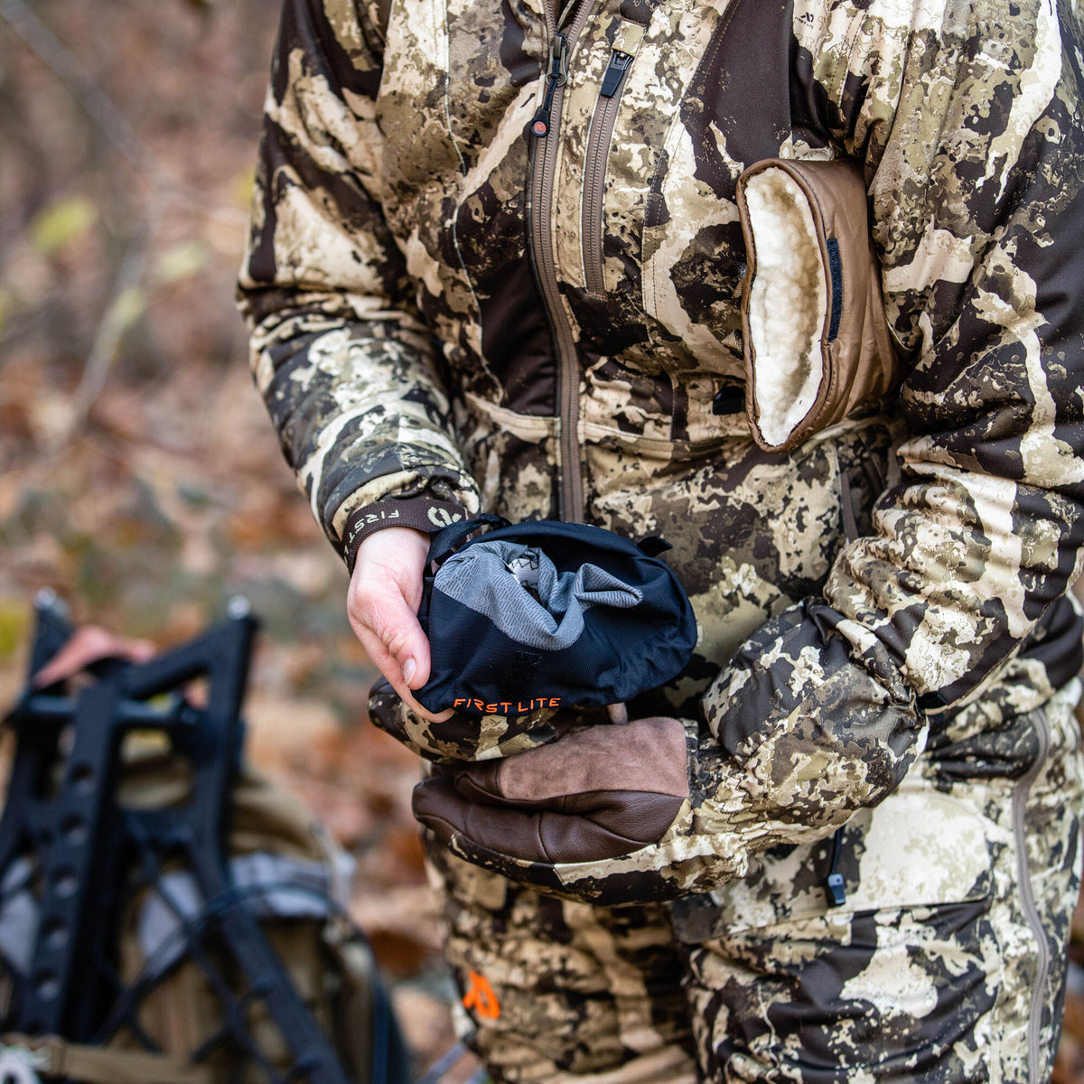 A person in First Lite camouflage holds a black stuff sack with a grey interior, wearing brown gloves, with a tan fleece-lined hand warmer visible on their waist.