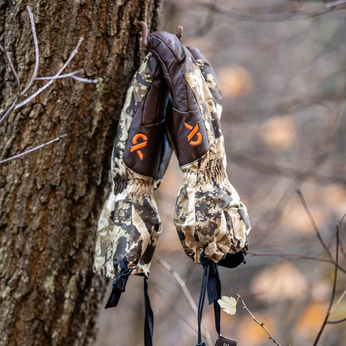 Pair of camouflage and dark brown leather gloves with orange logos hanging from a tree hook.