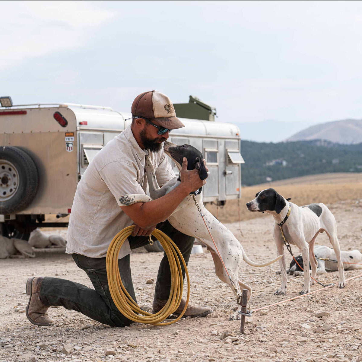 Man kneeling on the ground with a coiled rope, holding a dog, and another dog standing nearby in an outdoor setting.