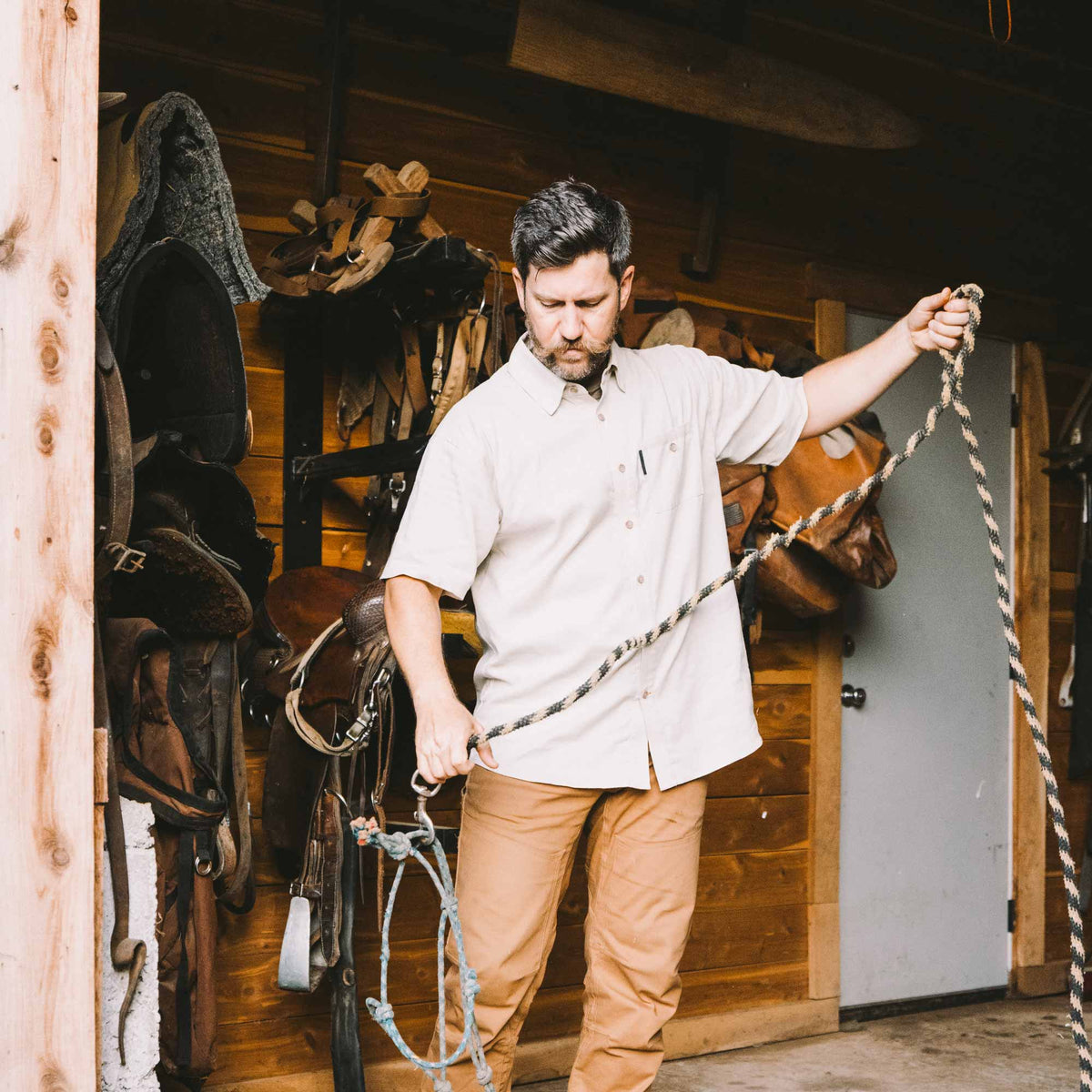 Man holding a rope in a stable with horse equipment in the background