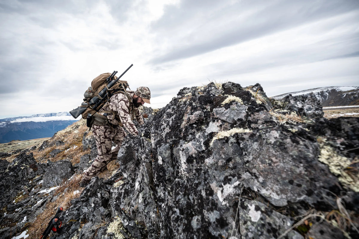 Hunter in tan camo outfit climbing rocky terrain with rifle & backpack.