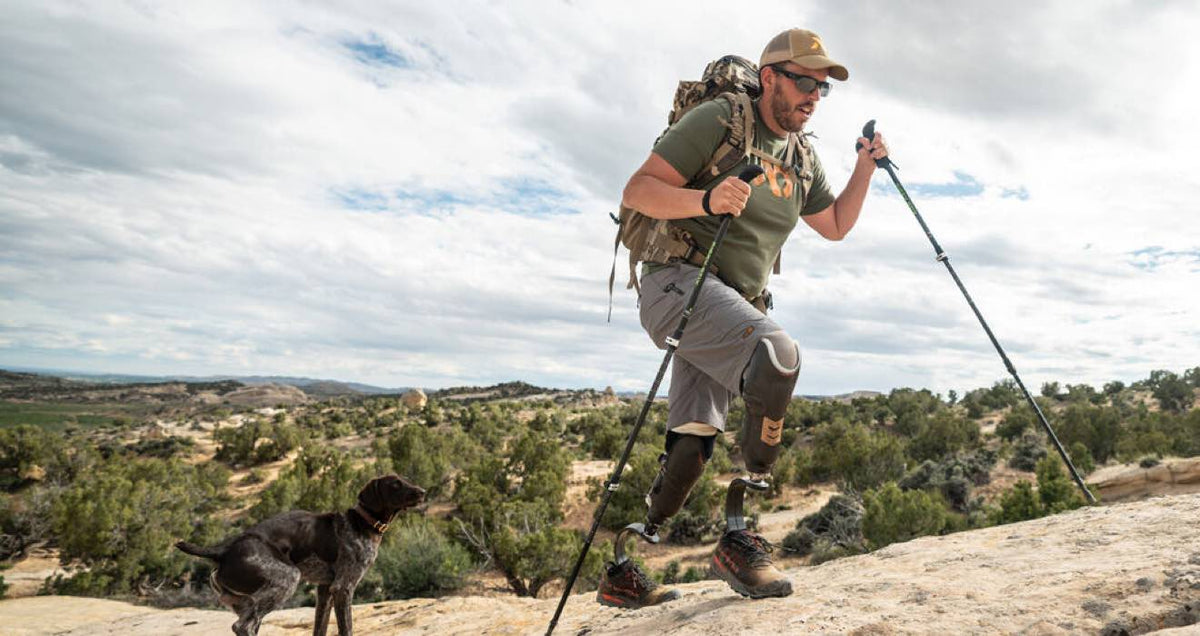 Man with dark bilateral prosthetic legs hikes uphill using poles.
