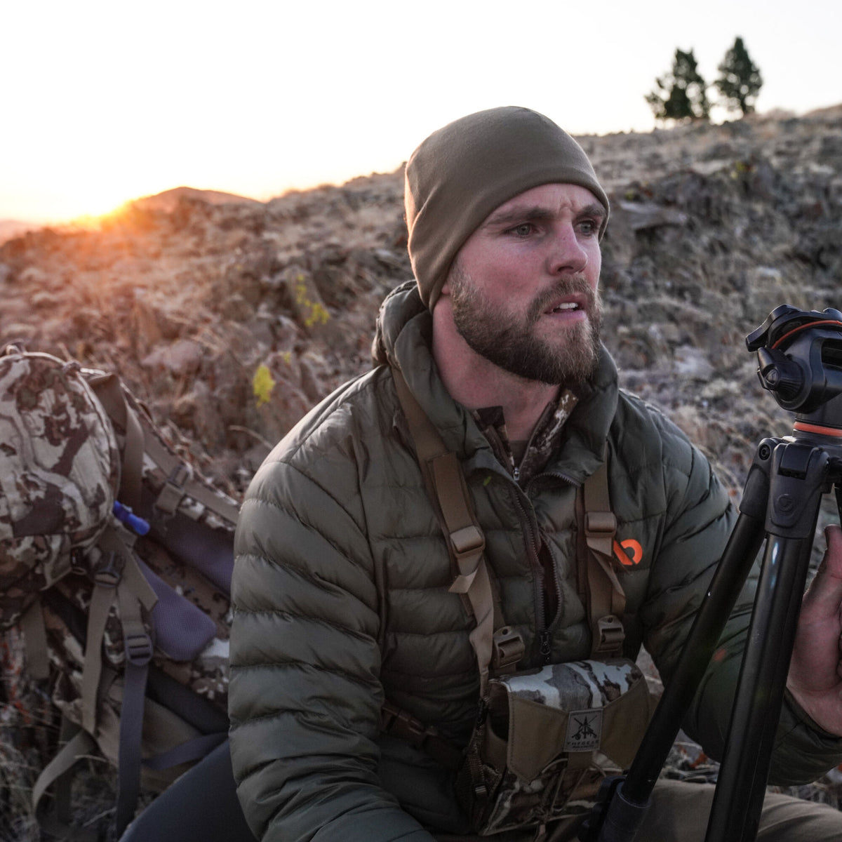 A man outdoors wears an olive green beanie and a quilted olive green puffer jacket with an orange chest logo.