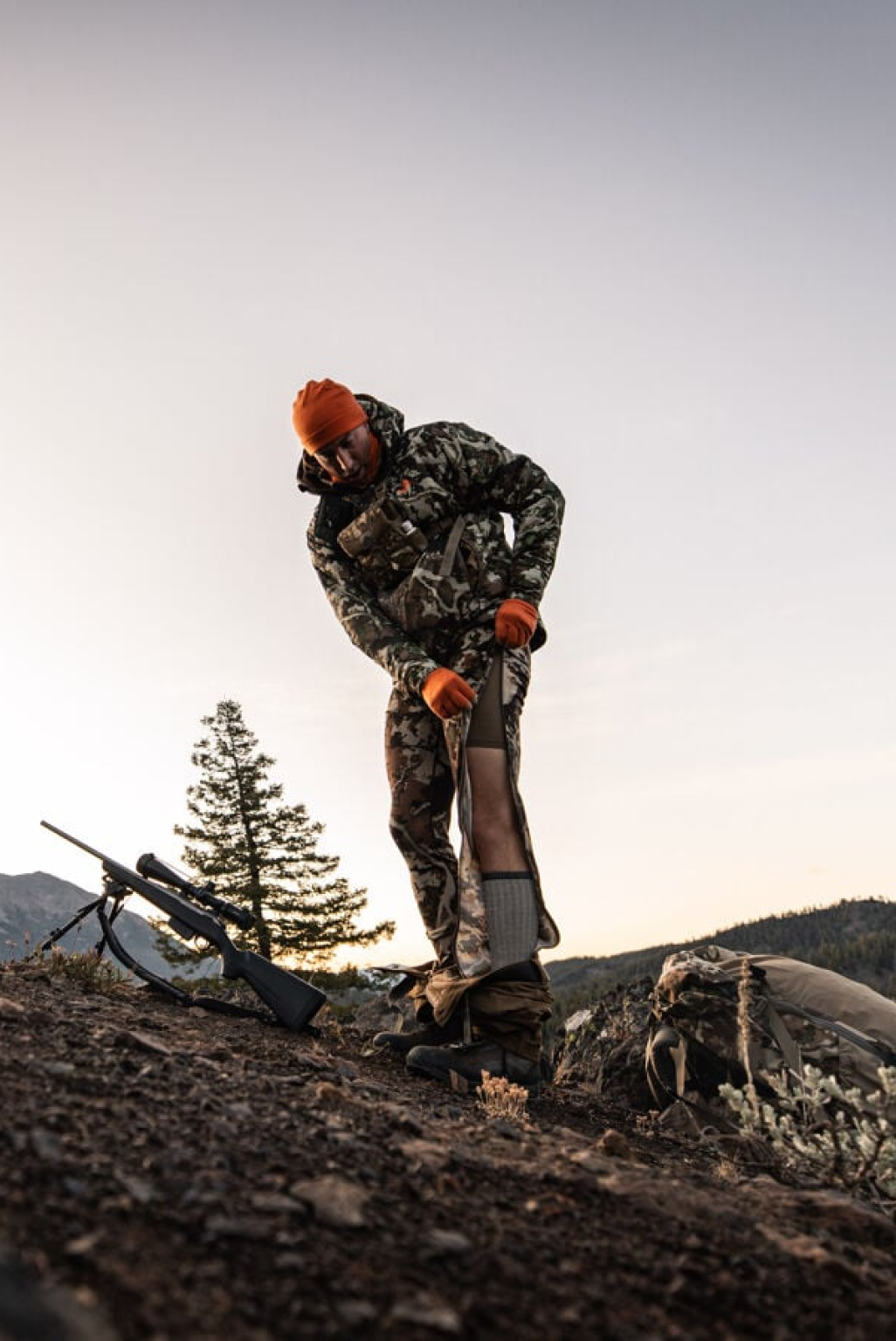 Camo hunting pants with full-length zippers being adjusted over a base layer.