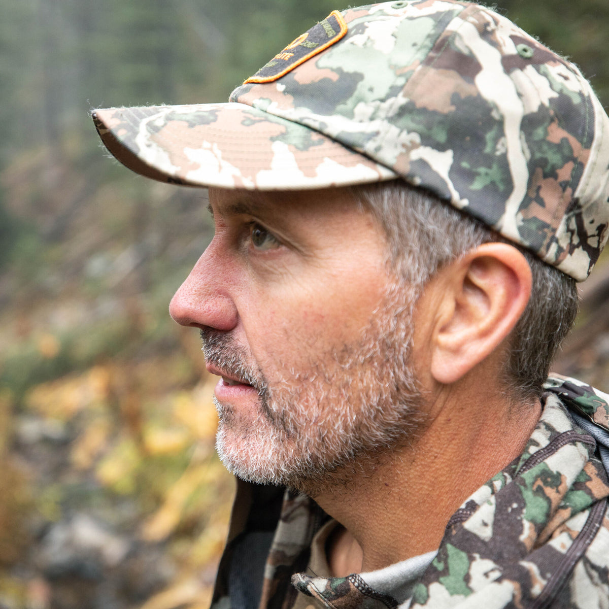Camouflage baseball cap with an orange-trimmed brand patch and ventilation eyelets, worn by a man.