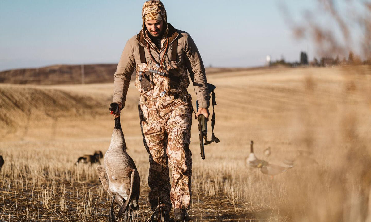 Hunter in brown camo bibs & jacket, holding a goose & shotgun in a field.