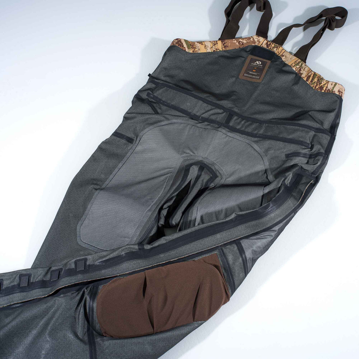 First Lite dark grey hunting bibs with camouflage top trim, brown suspenders, and brown knee pads.