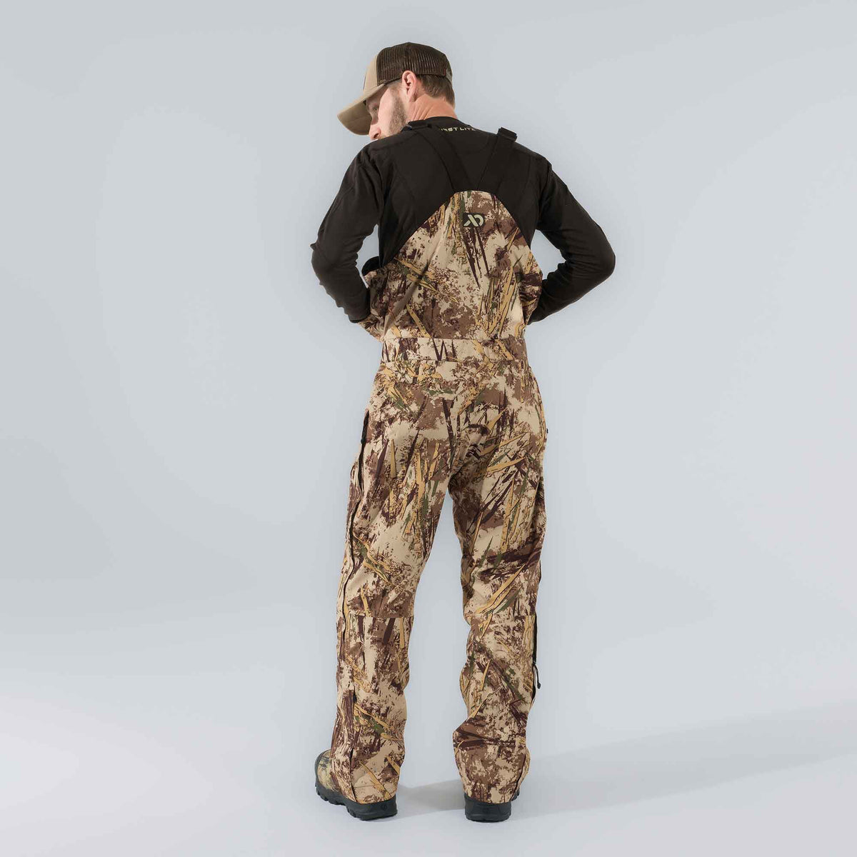 A man from behind wears brown and tan marshland camouflage hunting bibs with adjustable shoulder straps and a logo on the bib.