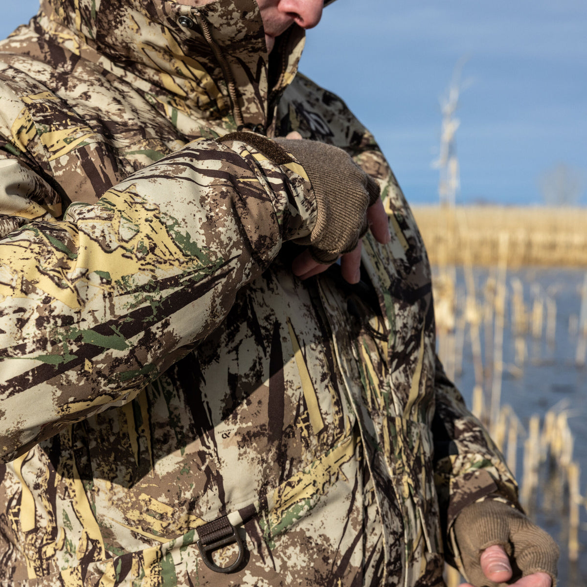 Brown, tan, and green camouflage hunting jacket with a D-ring at the hem, worn by a person in fingerless gloves by a marsh.