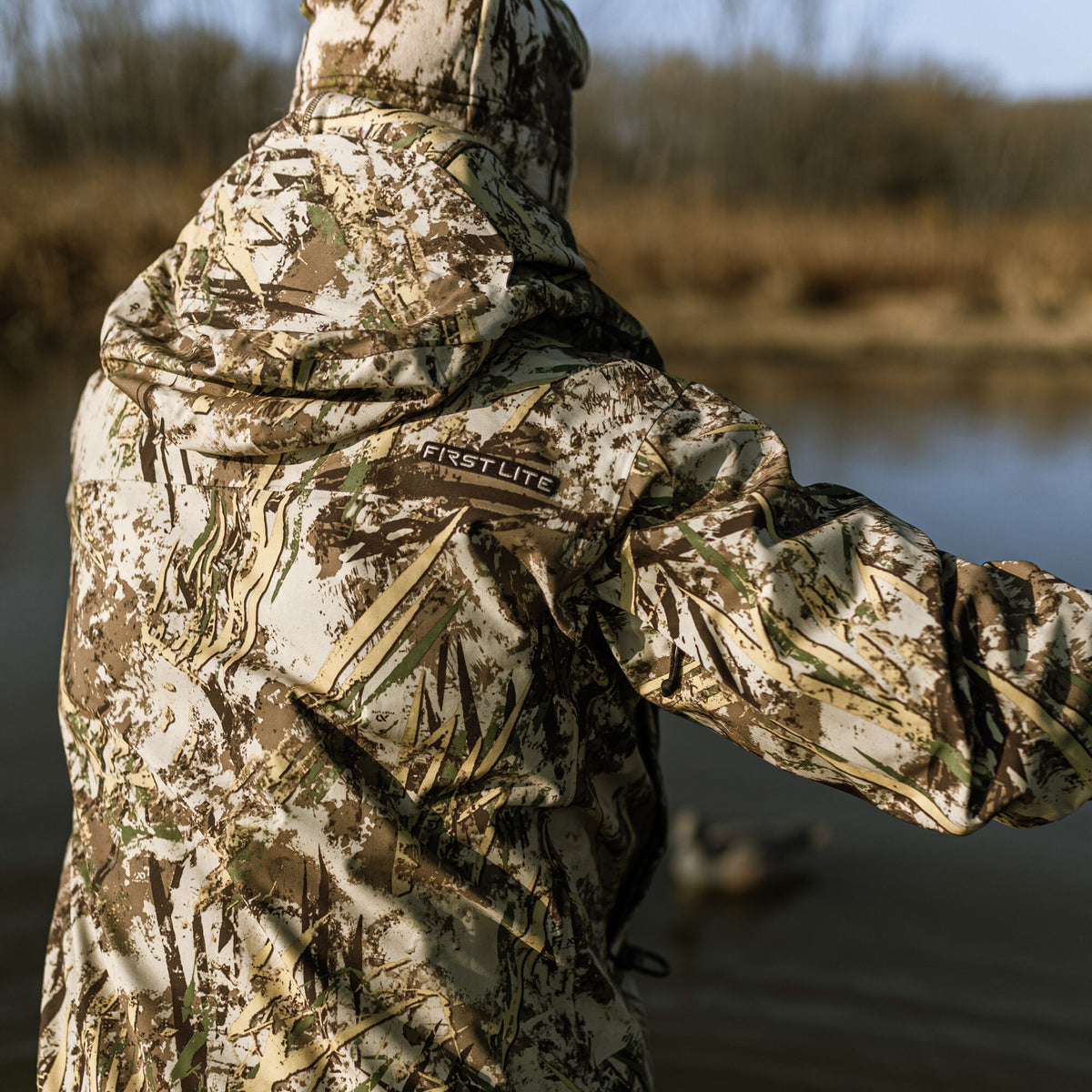 Hooded First Lite camouflage jacket in a tan, brown, and green reed pattern, viewed from the back.