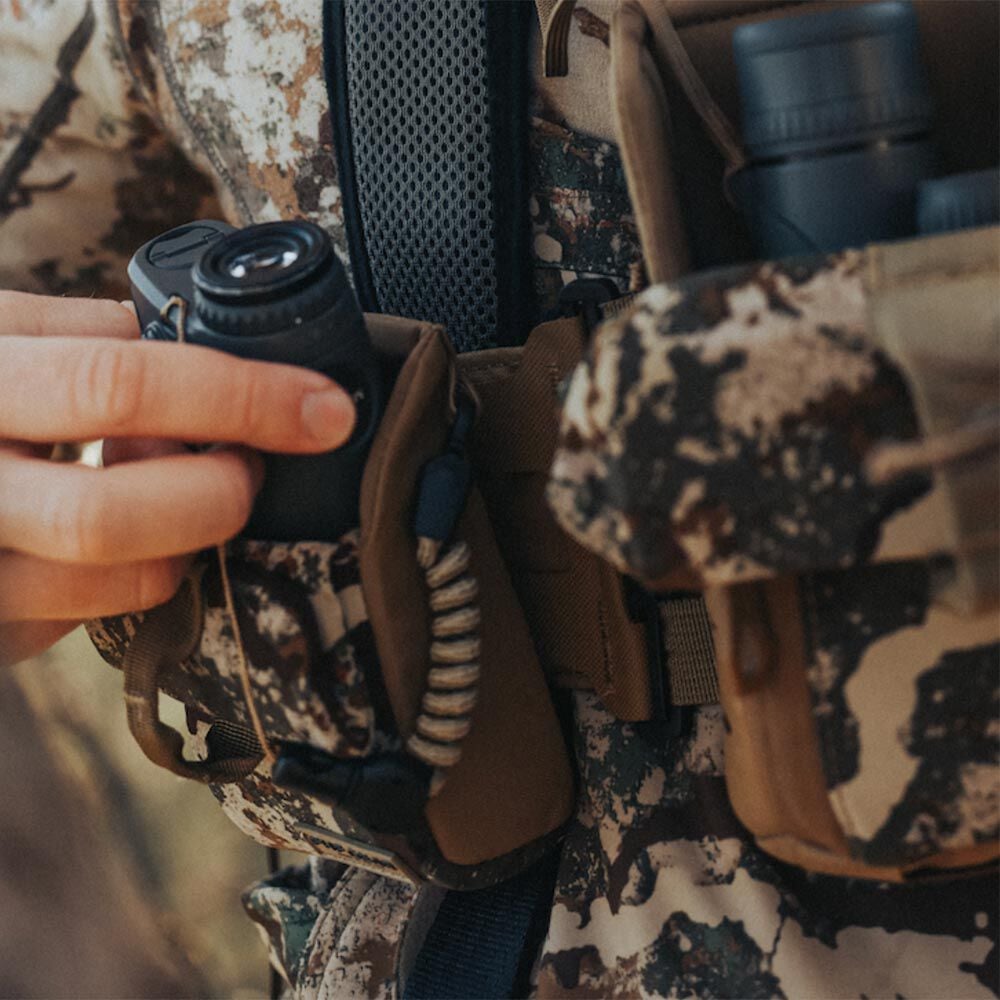 A hand inserts a black rangefinder into a tan and camouflage chest harness featuring multiple pouches, binoculars, and a coiled lanyard.