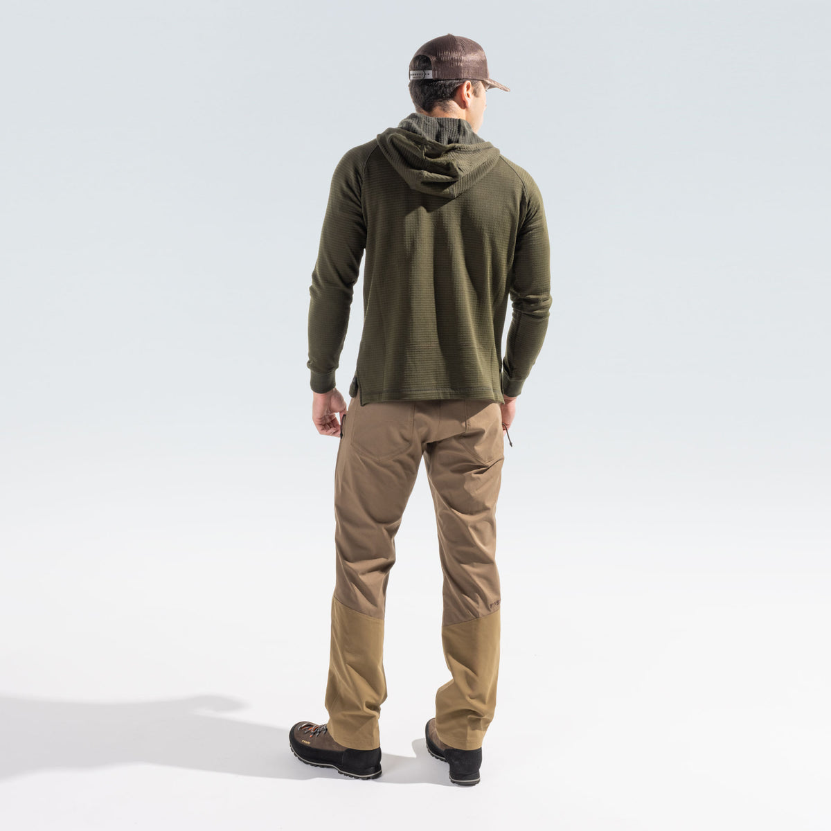 Man wearing tan brush pants and a green grid hoody, standing facing away from the camera on a white background.