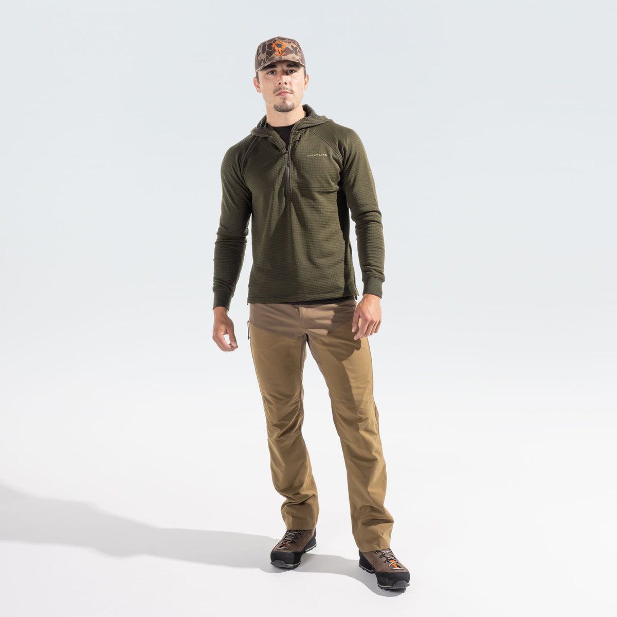 A man modeling an olive green quarter-zip hooded pullover, tan pants, and a camouflage cap against a light gray studio background.