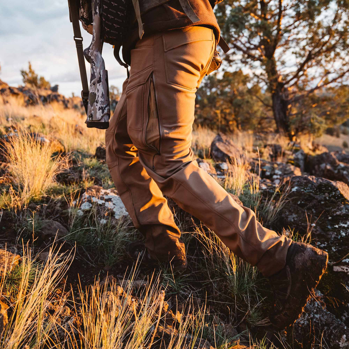 Person wears tan outdoor pants featuring zippered thigh pockets while hiking in a rocky, grassy landscape with a rifle.