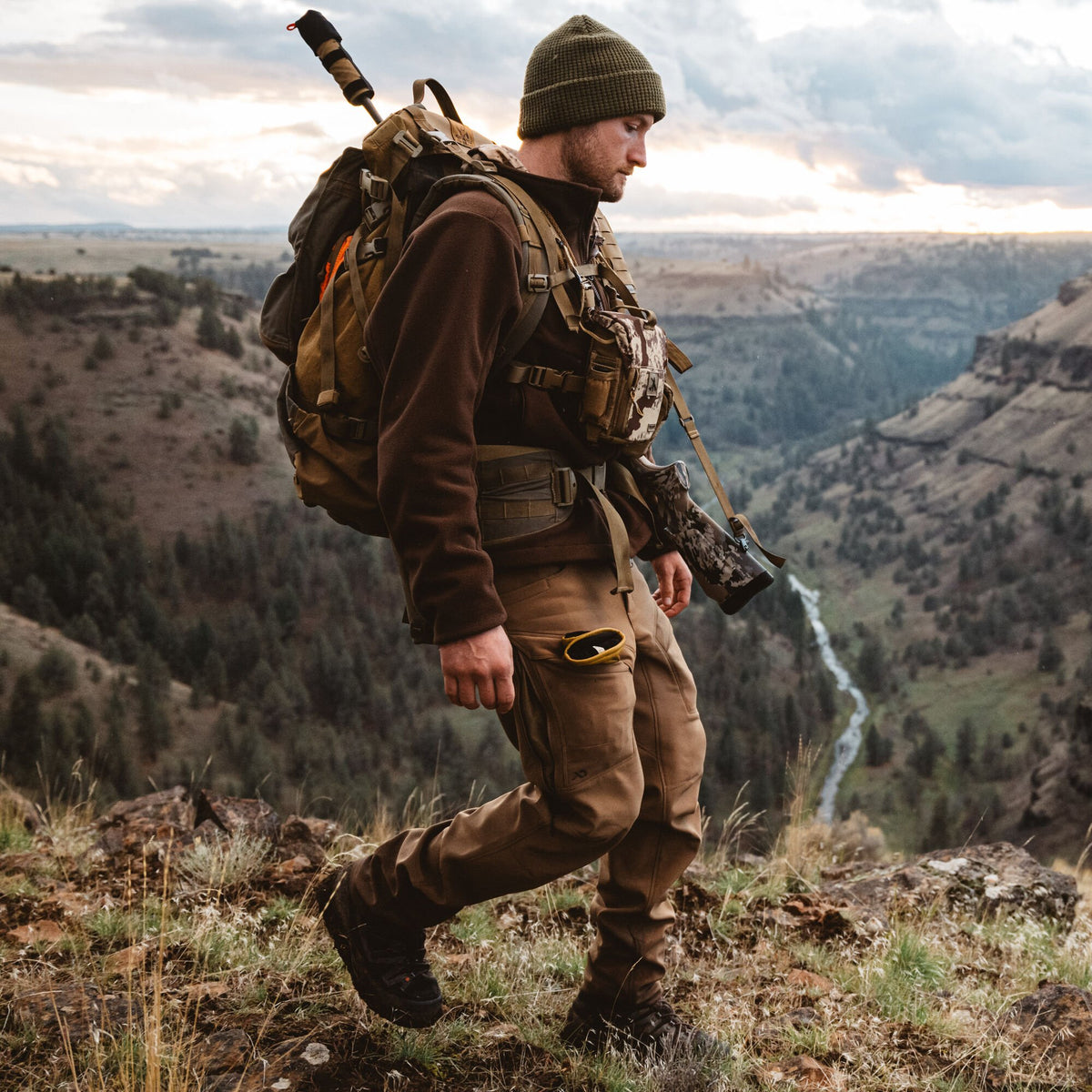 Brown First Lite hiking pants featuring a side leg zipper and thigh pocket, worn by a man walking outdoors.