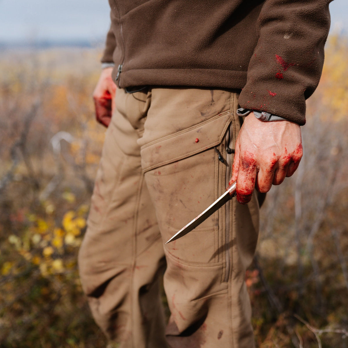 A person wears a blood-stained brown fleece jacket and tan cargo pants, holding a bloody silver knife.