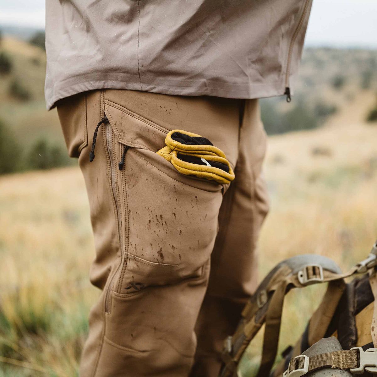 Tan pants with dirt splatters, featuring zippered pockets and yellow work gloves in an open side pocket.