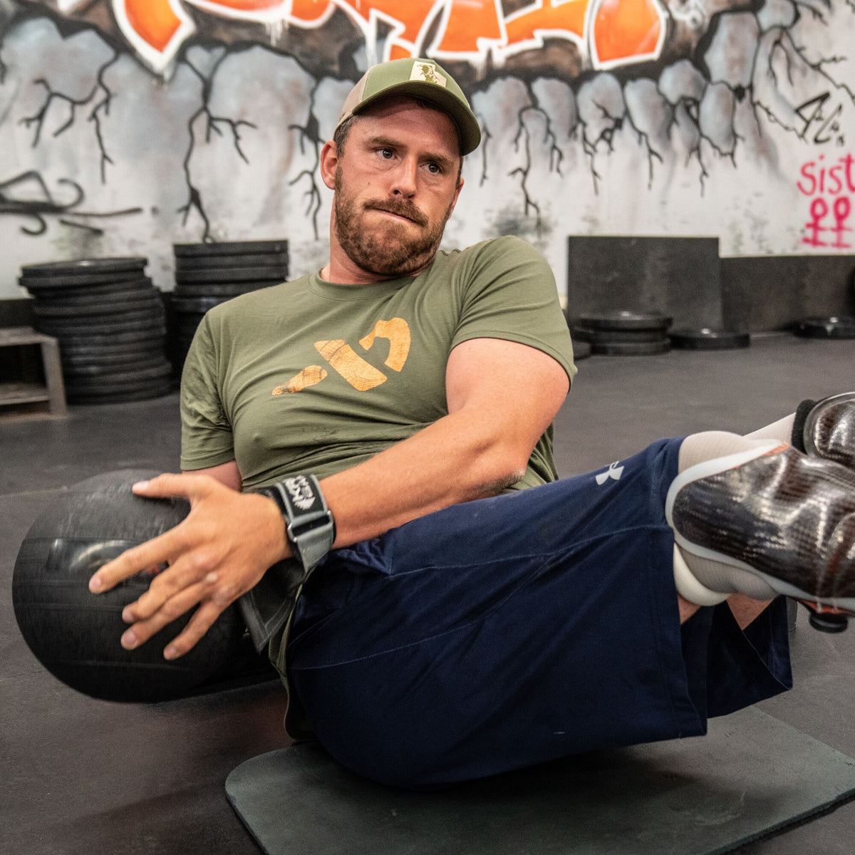 A man in an olive green t-shirt with an orange axe graphic, an olive green and tan hat, and dark blue shorts performs a seated Russian twist with a black medicine ball.