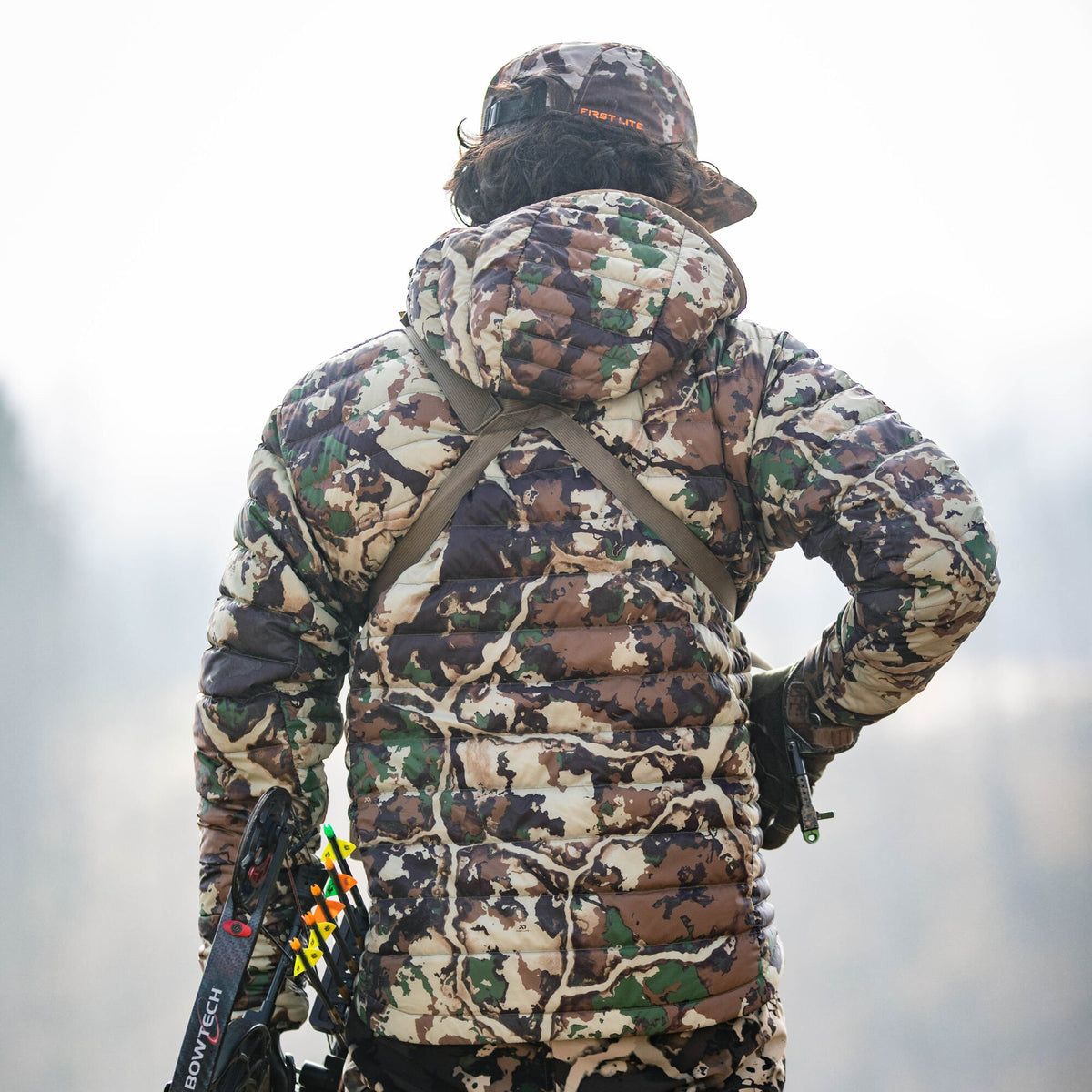A person viewed from behind wears a First Lite Fusion camouflage quilted puffer jacket and hat, holding a Bowtech compound bow with arrows.