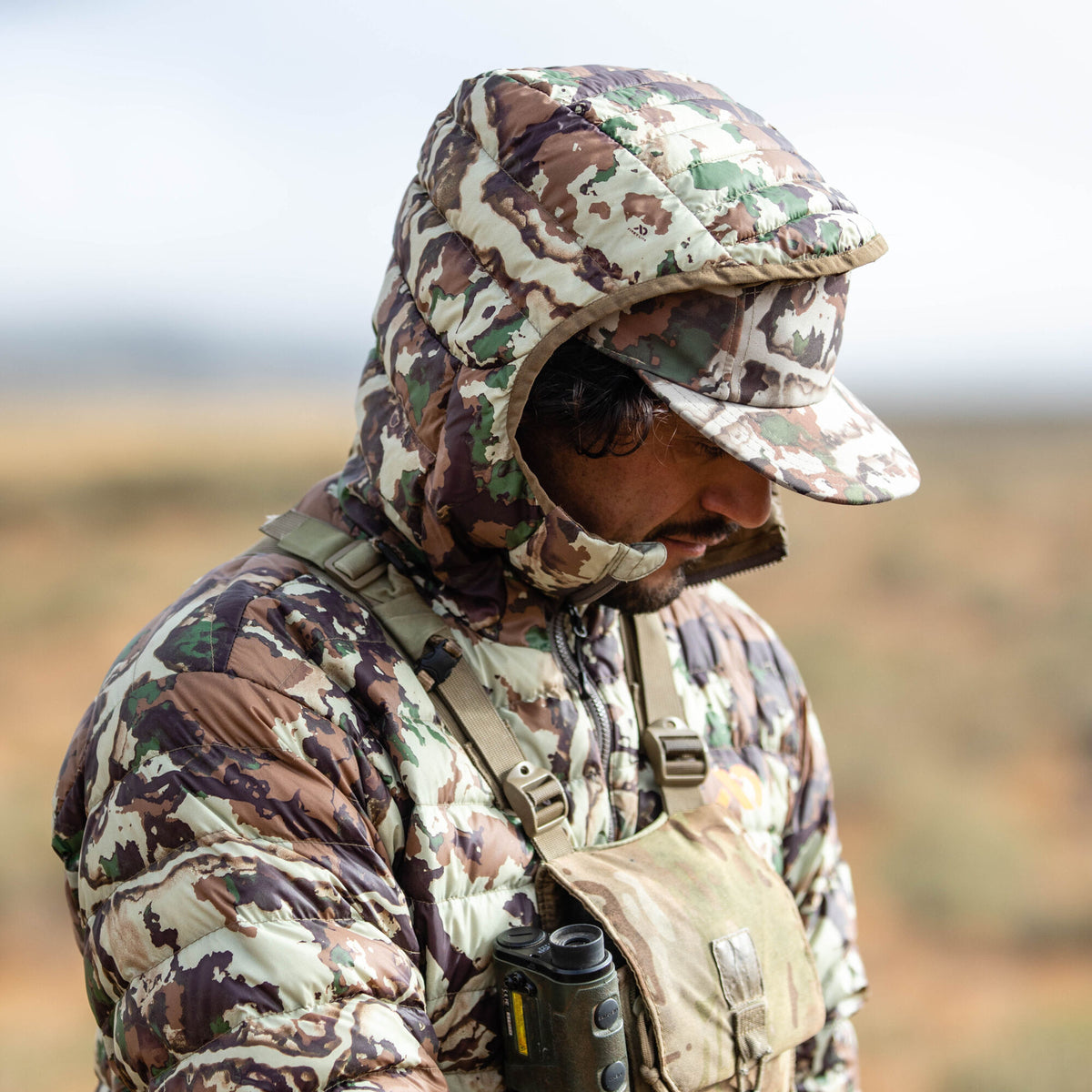 A man wears a quilted, hooded camouflage jacket and matching baseball cap with a rangefinder in a chest harness.