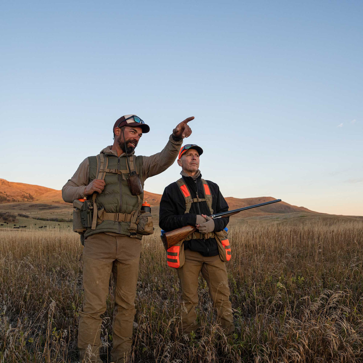 Two men wearing upland vests and on a bird hunt, standing on the prairie wearing upland hunting clothing.