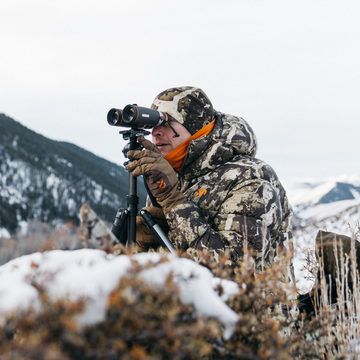 Black binoculars with orange accents mounted on a tripod, being used by a person in a snowy mountain landscape.