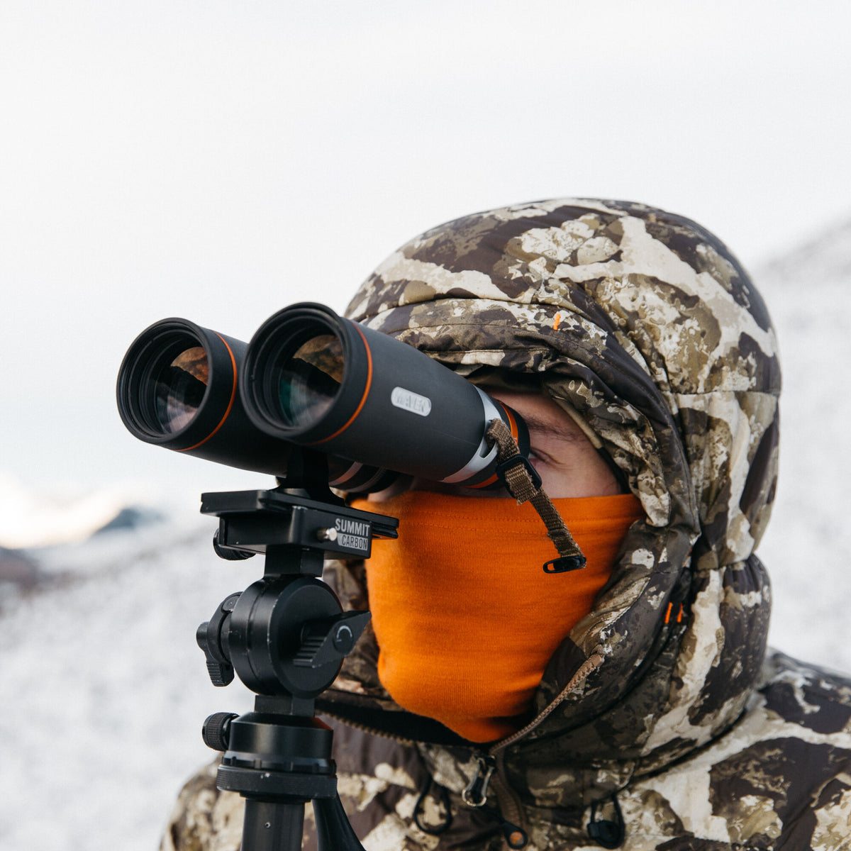 Black Maven binoculars with orange trim, mounted on a Summit Carbon tripod, in use by a person in a camo jacket and orange gaiter.