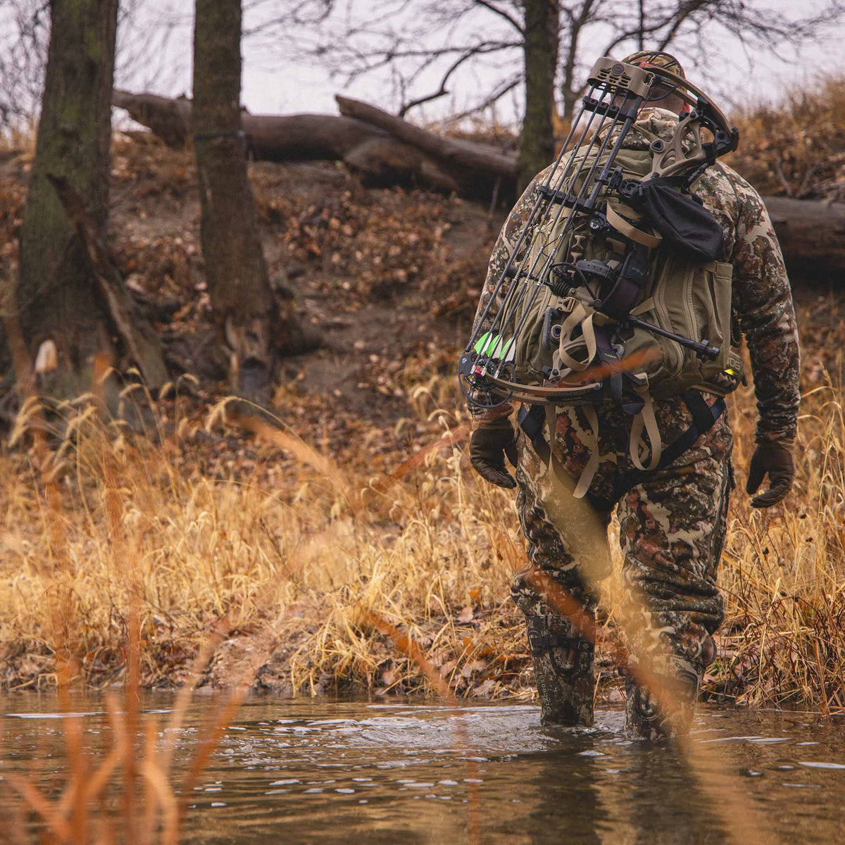 A hunter in brown and green camouflage, wearing a harness and carrying a tan compound bow with green-fletched arrows and an olive backpack, wades through a shallow stream.