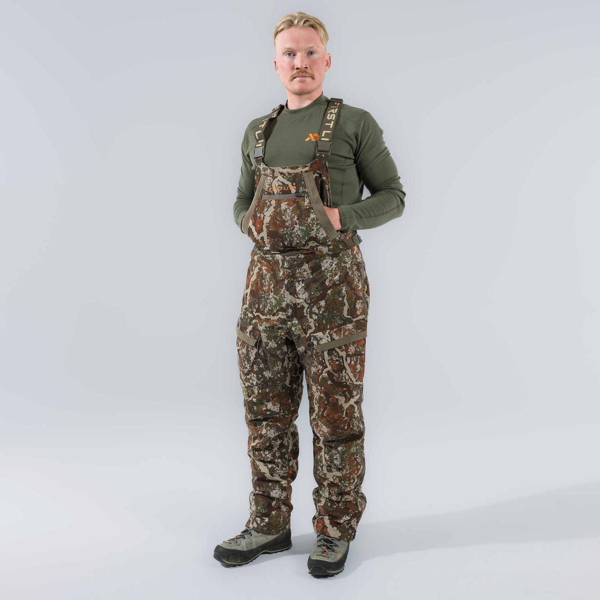 Man in camouflage hunting bib overalls with cargo pockets and First Lite branded straps.