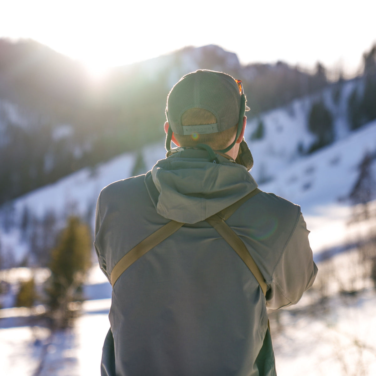 Gray hooded jacket with a khaki chest harness, and an olive green mesh-back cap with sunglasses resting on top, viewed from behind in a snowy mountain landscape.