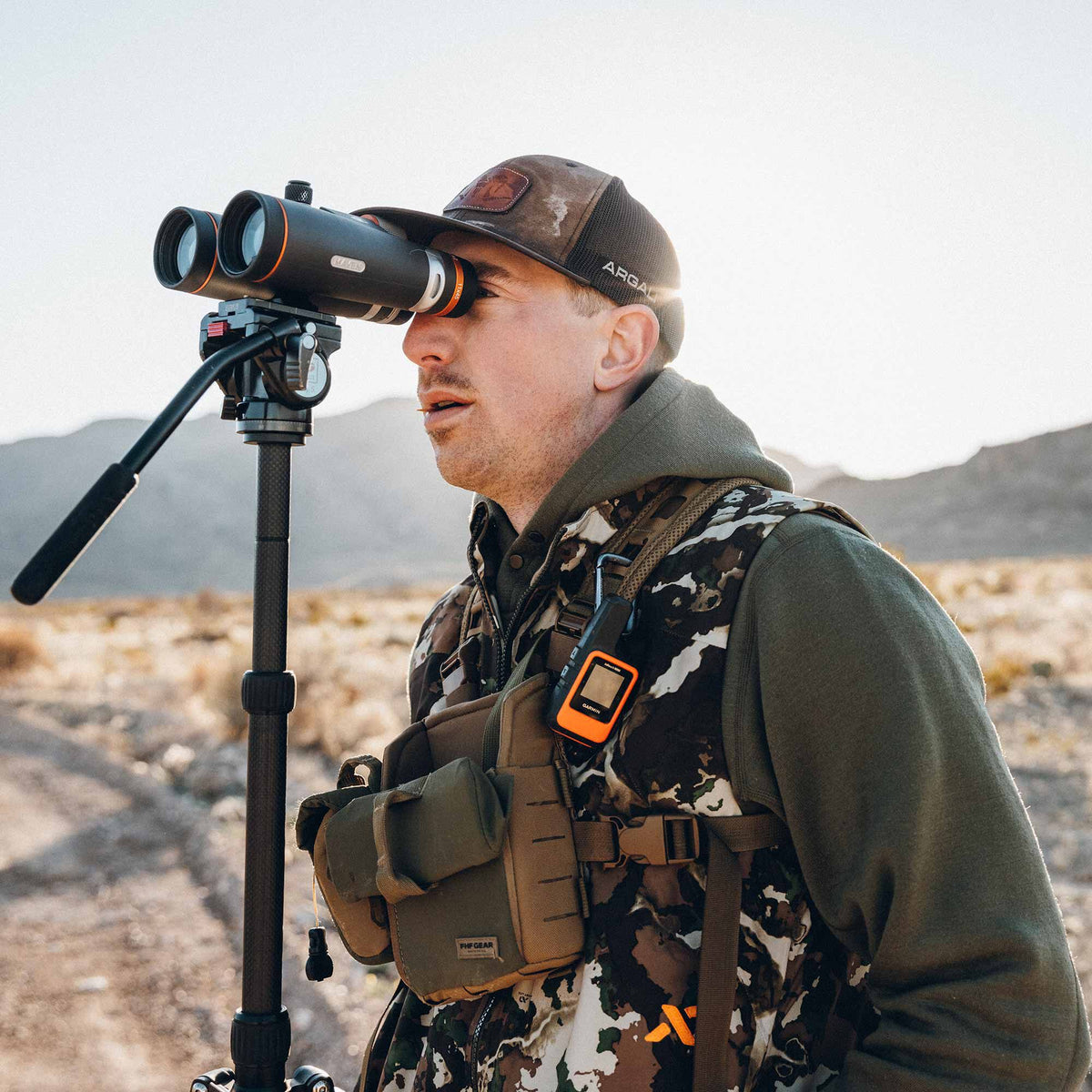Black Maven binoculars with orange lens rings, mounted on a carbon fiber tripod, being used by a person wearing a camouflage vest.