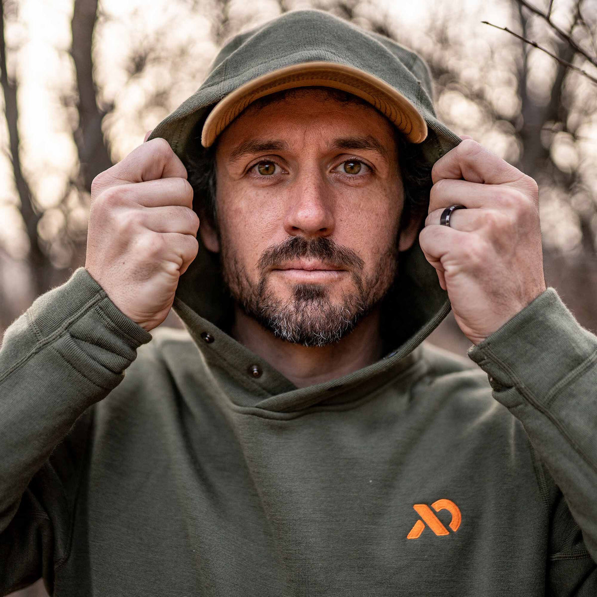 Olive green hoodie with a snap placket, ribbed cuffs, and an orange chest logo, worn with the hood up over a tan cap.