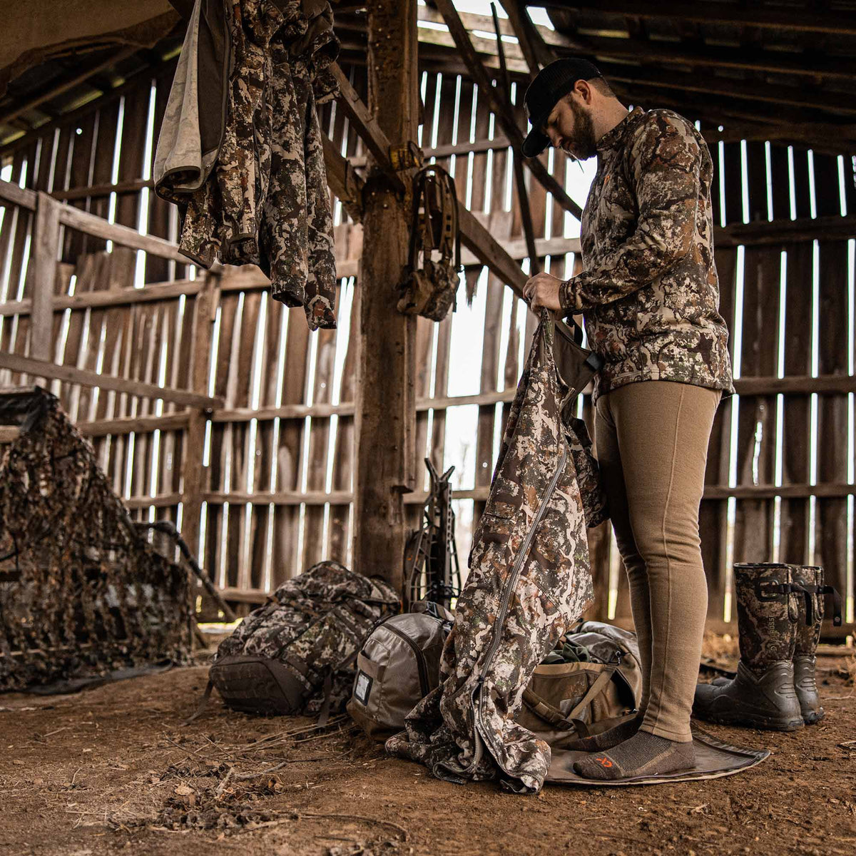 Man inspects brown and green camouflage hunting outerwear with full-length zippers inside a rustic barn filled with hunting equipment.