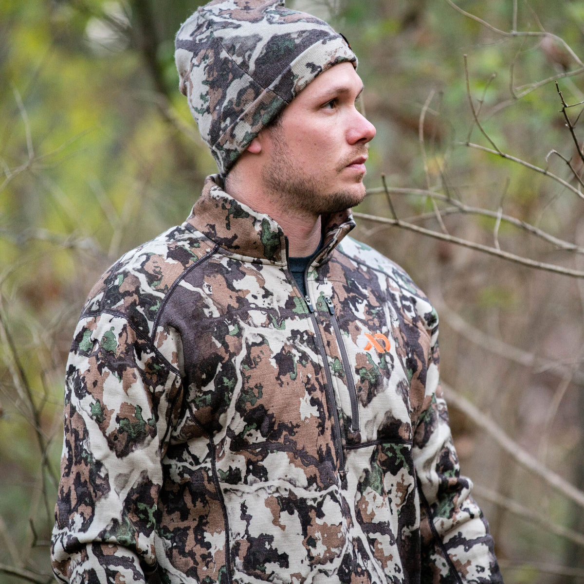 Man wearing a brown, green, and white camouflage quarter-zip jacket with an orange chest logo, a zippered pocket, and a matching beanie.