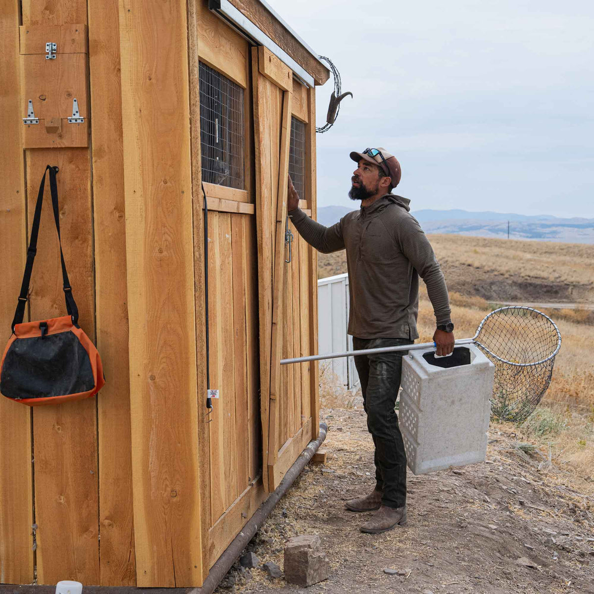 Man with a beard and cap holding a net and container near a wooden shed in a rural setting.