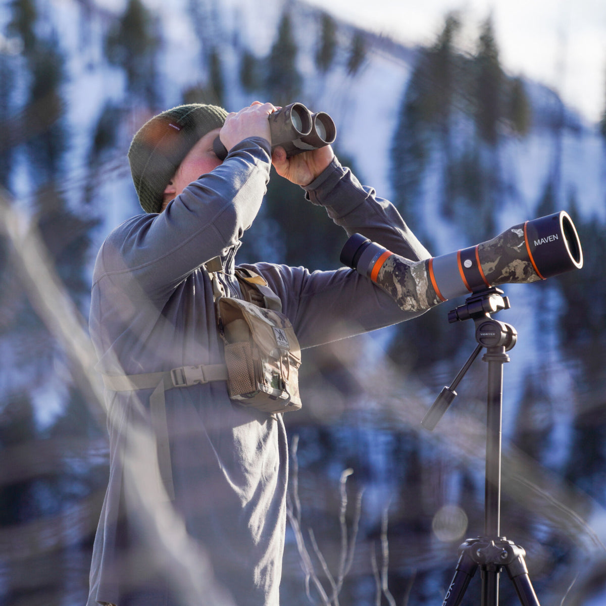 A person views a snowy mountain through dark binoculars, with a black, gray, and camouflage Maven spotting scope featuring orange bands set on a Vortex tripod nearby.