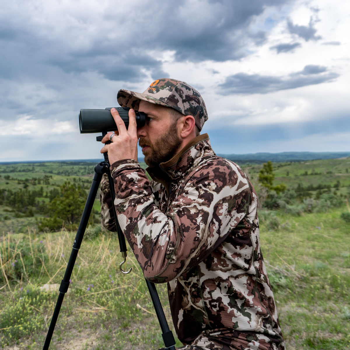 A dark green spotting scope mounted on a black tripod is being used by a man wearing camouflage.