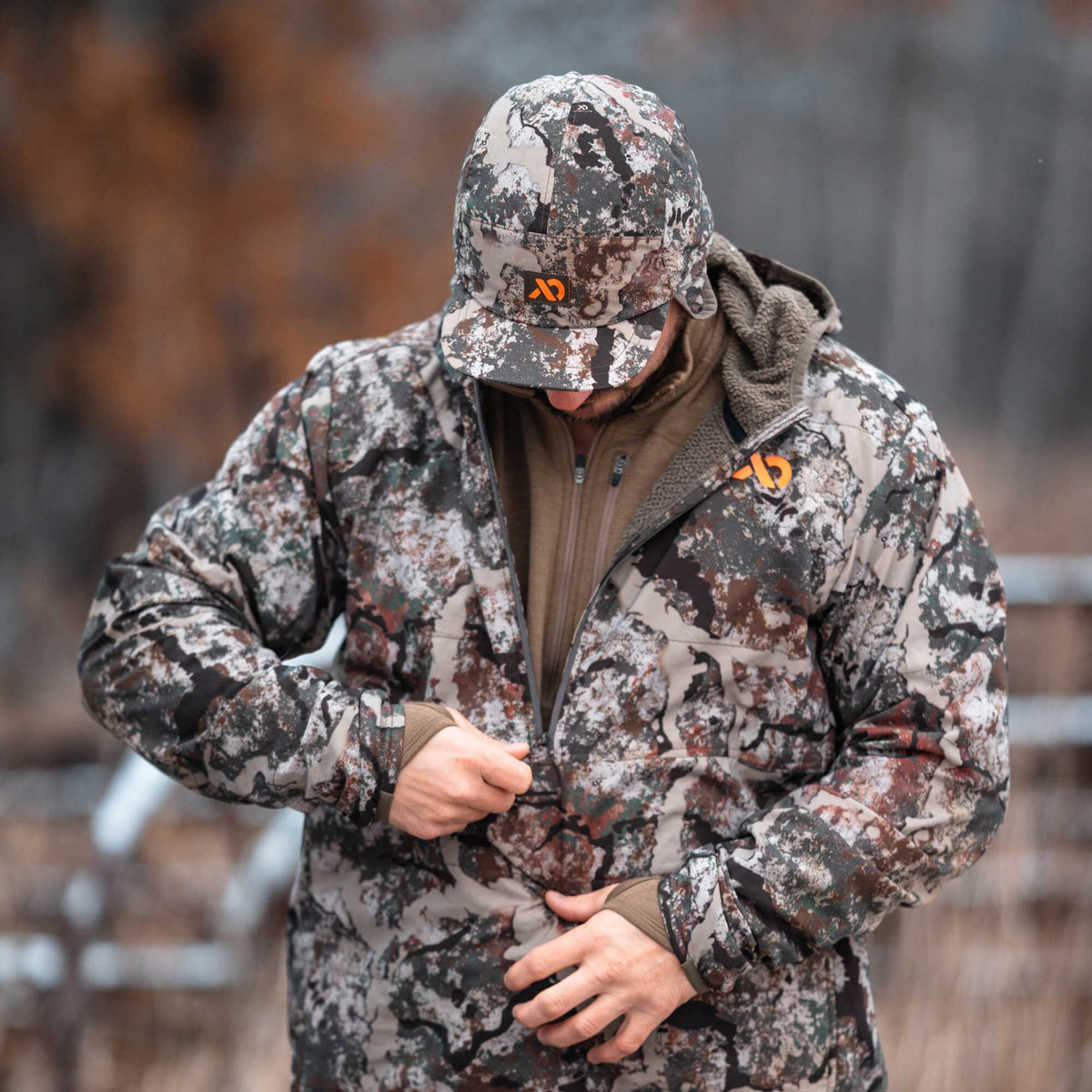 A man in a grey, white, brown, and green camouflage hunting jacket and cap, both with orange logos, is zipping up his jacket.