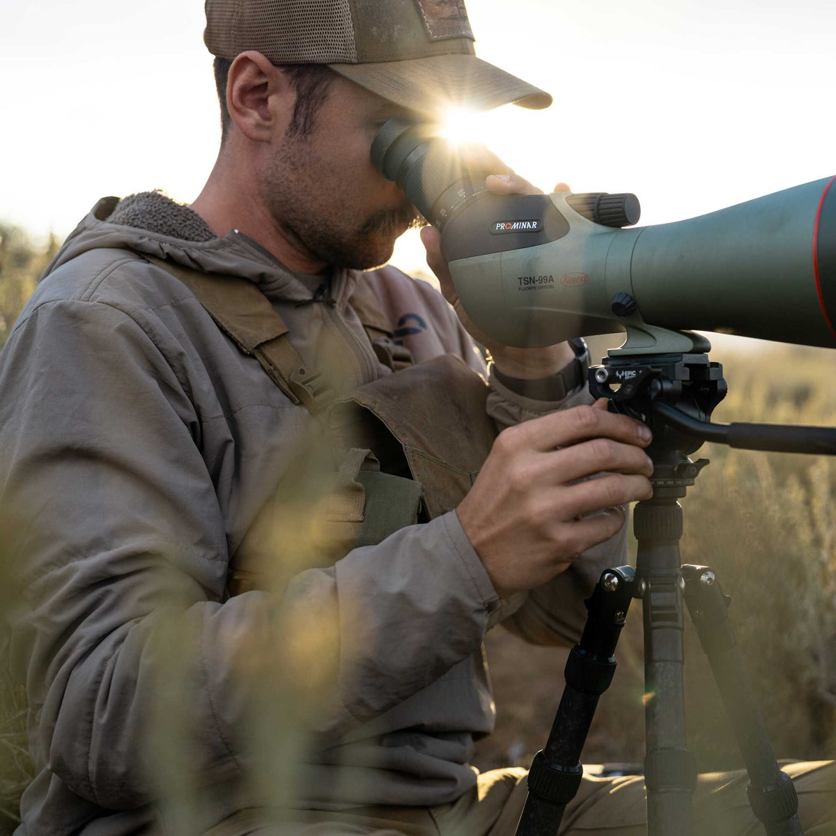 A man uses an olive green Kowa TSN-99A Prominar Fluorite Crystal spotting scope mounted on a black tripod outdoors.