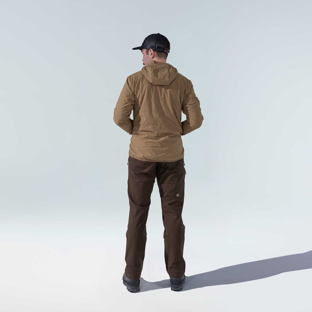 Rear view of a man wearing a tan hooded insulated jacket, dark brown pants, and a black mesh-back cap.